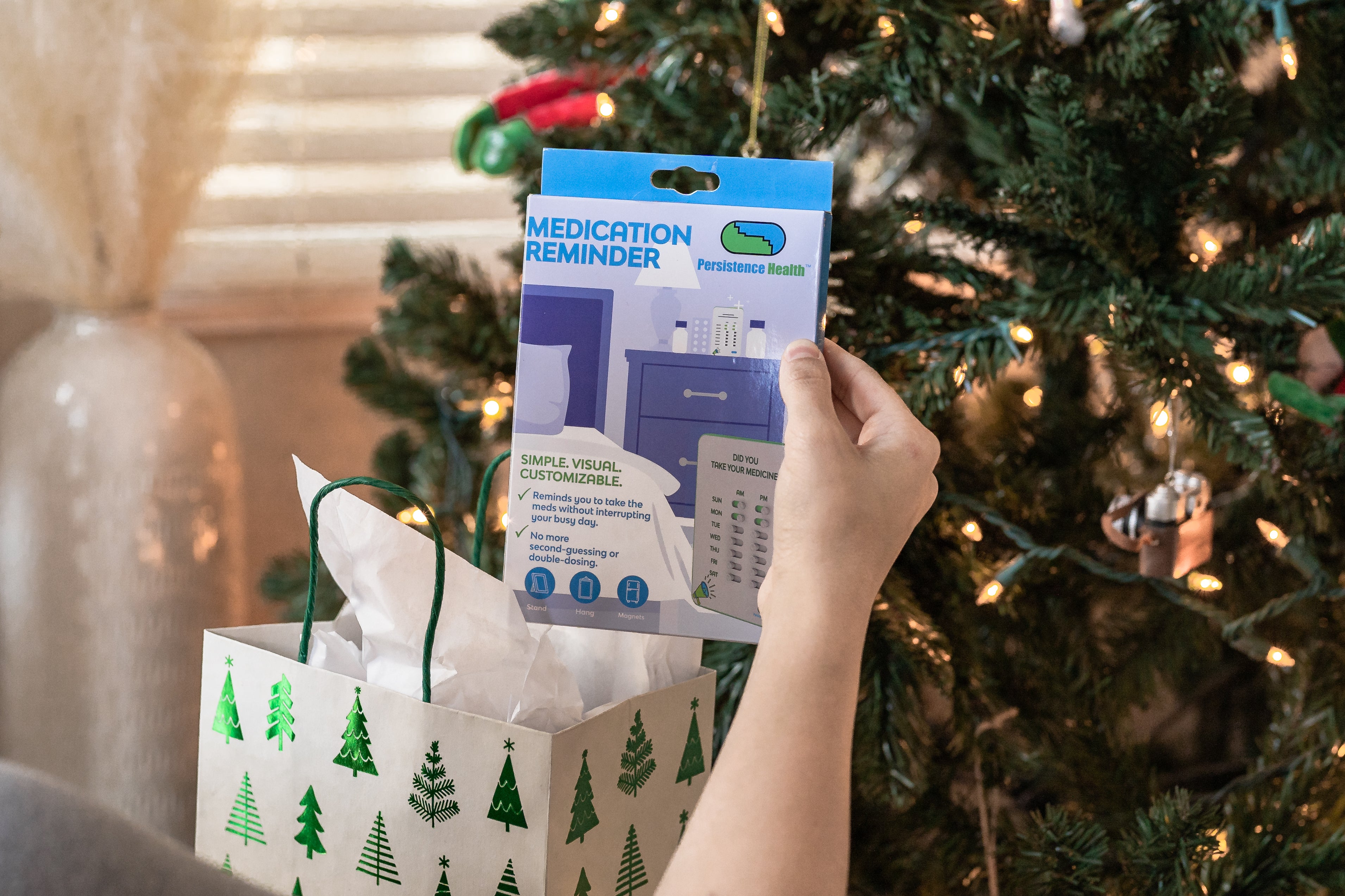 Hand holding a medication reminder device package in front of a Christmas tree