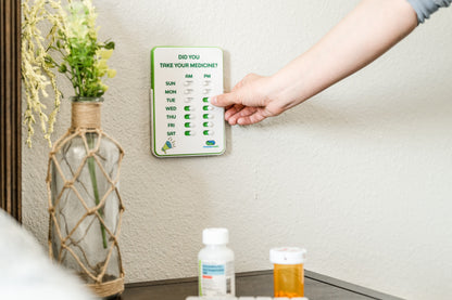 Hand interacting with a digital pill reminder device on a nightstand next to a vase with plants and medicine bottles.