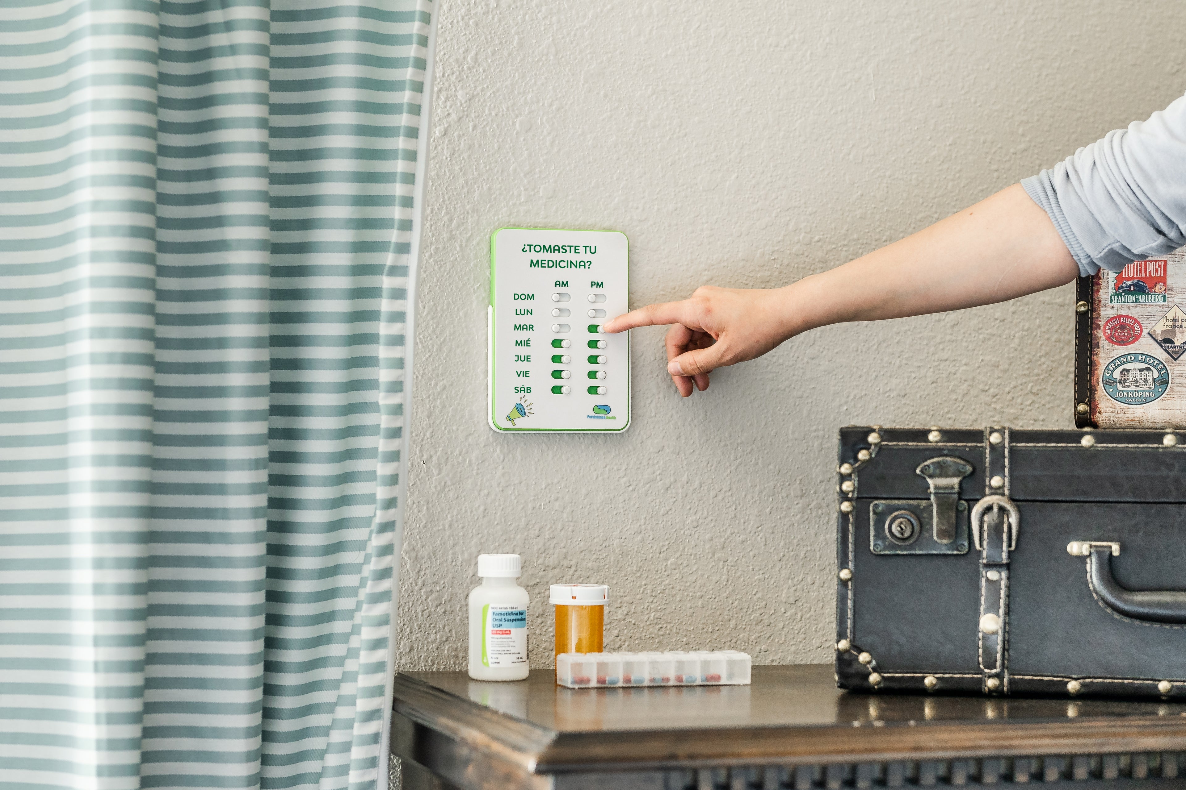 Person pointing at a wall-mounted digital thermometer next to a suitcase and medical supplies.
