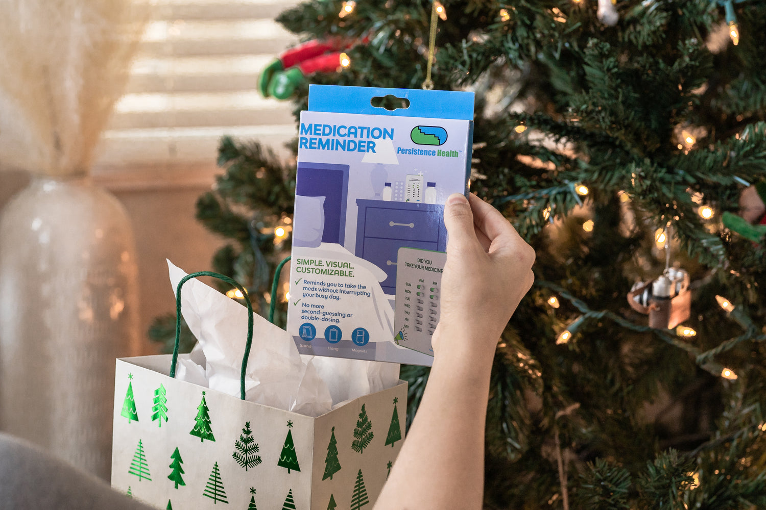 Hand holding a medication reminder device package in front of a Christmas tree