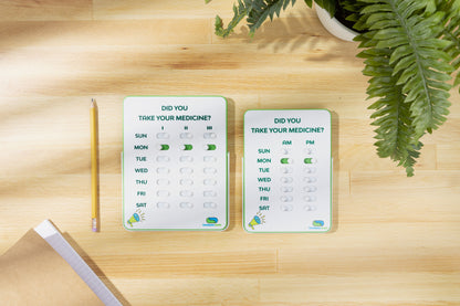 Two pill reminder cards on a wooden surface with a plant in the background