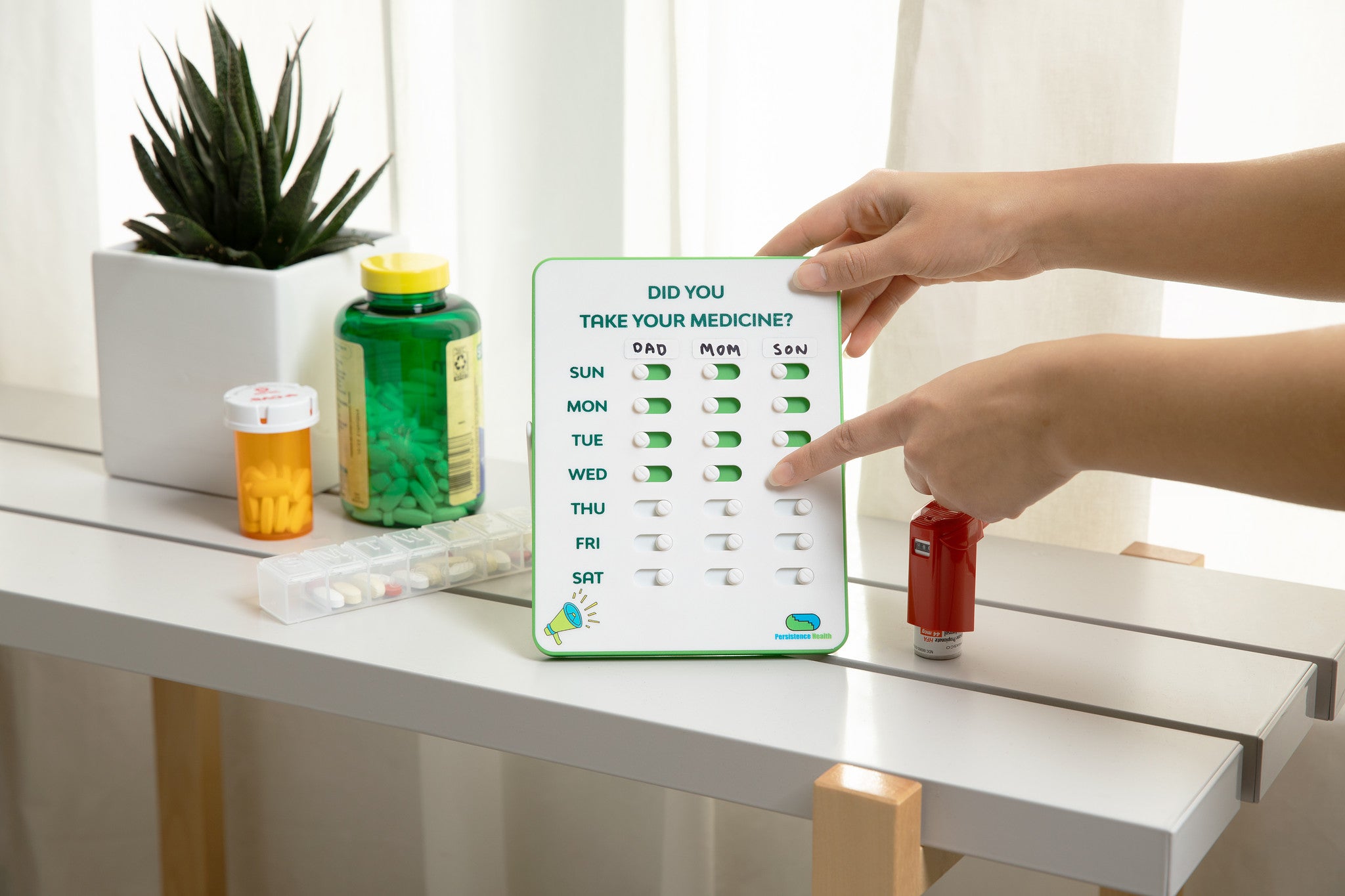 Hand interacting with a digital pill reminder on a table with medications and a plant.