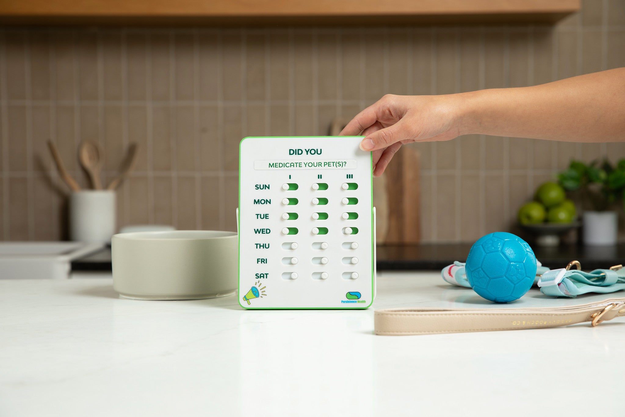 Hand holding a pet medication reminder card on a kitchen counter with a blue ball and pet toys.